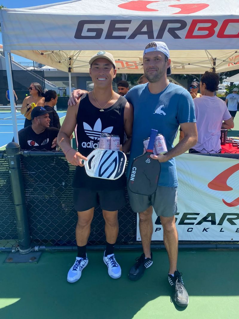 Michael Kuhlman, on the right in a backwards cap, with another player under the event tent at a pickleball tournament, both holding paddles