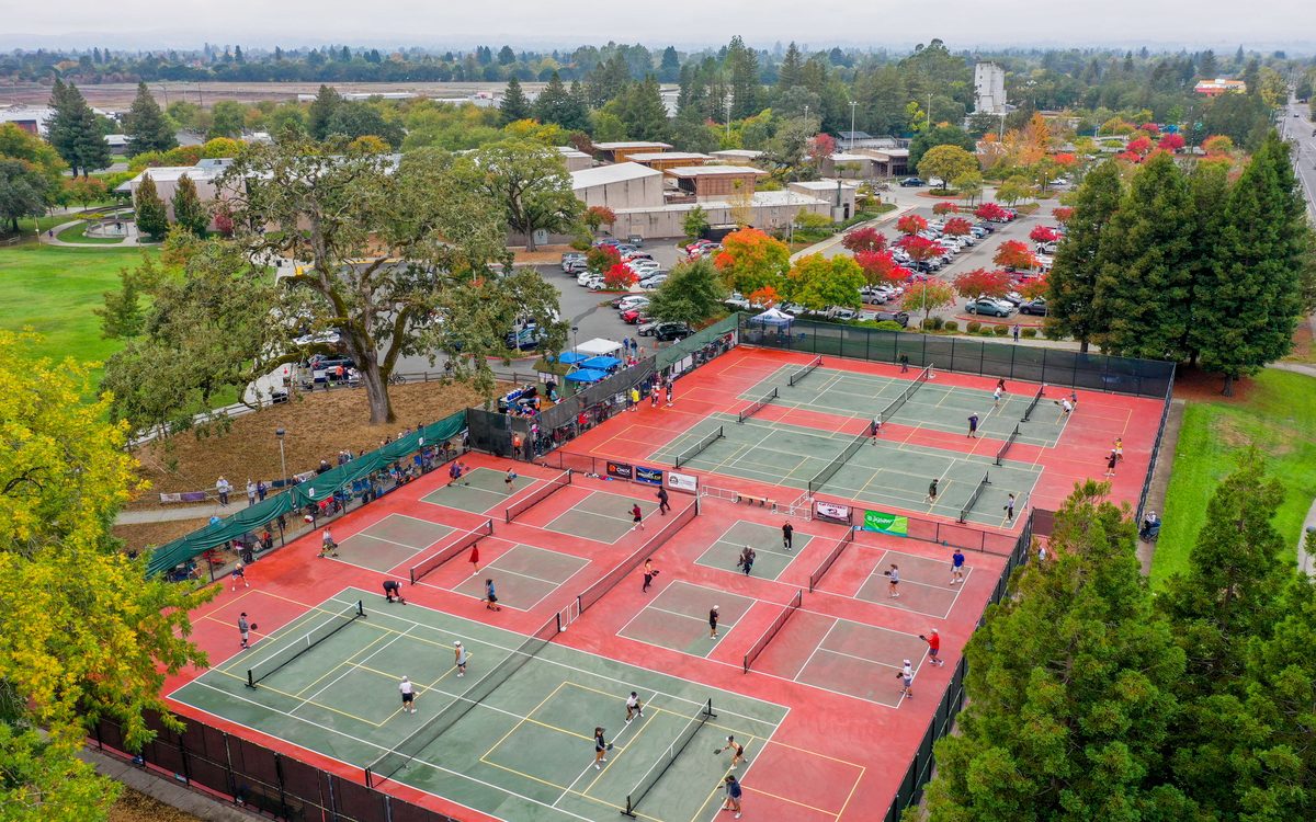 Aerial view of the twelve green-and-maroon pickleball courts at Finley Community Park, Santa Rosa, in full play during a tournament, with the community center and oaks behind