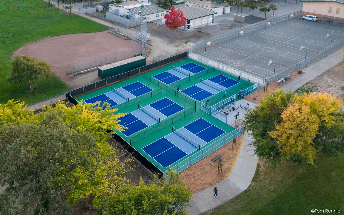 Aerial view at sunset of the six blue and green pickleball courts at Hiram Lewis Park, Windsor, with players on court and the Russian River valley beyond